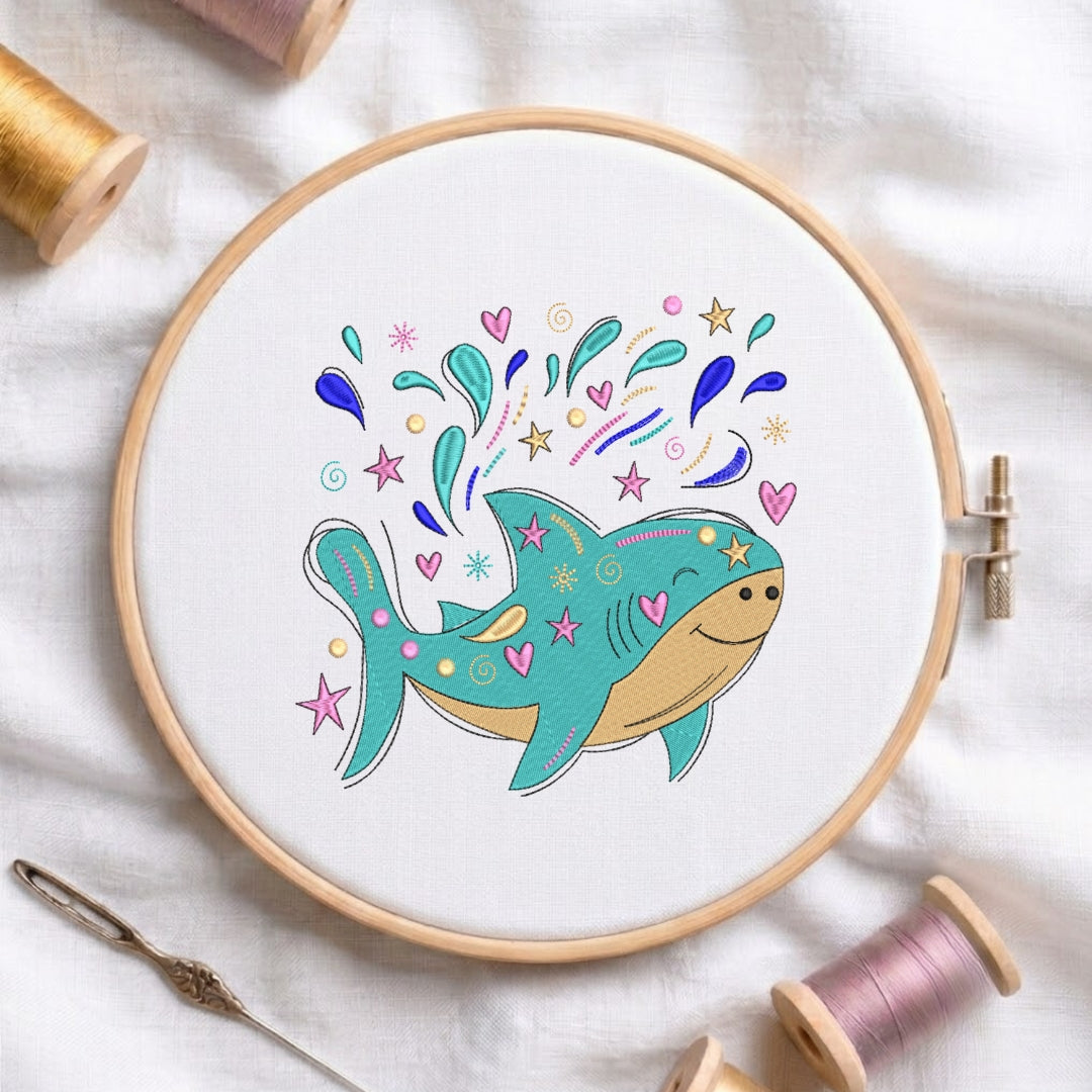 A circular embroidery hoop holding white fabric with a colorful cartoon fish design in progress. The fish is teal and beige with pink hearts and blue, purple, and yellow decorative splashes around it.