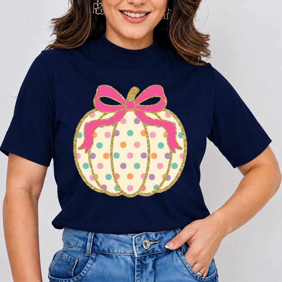 A woman wearing a navy blue t-shirt with a large graphic of a pumpkin decorated with colorful polka dots and a pink bow with gold glitter trim, paired with blue jeans.