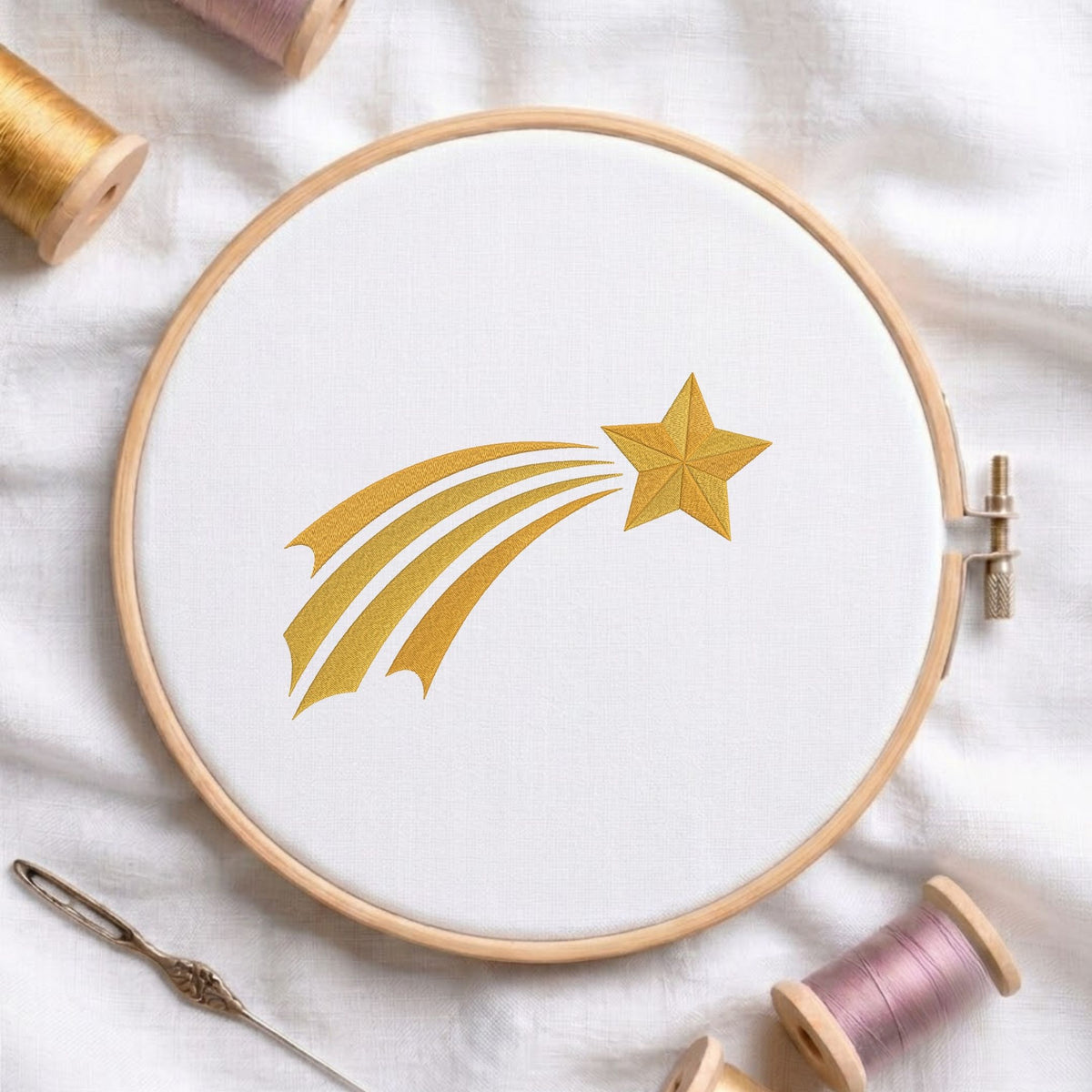 A circular embroidery hoop holding white fabric with a metallic gold embroidered shooting star design. The hoop is styled on a white fabric background with spools of thread, a needle, and a pin around it.