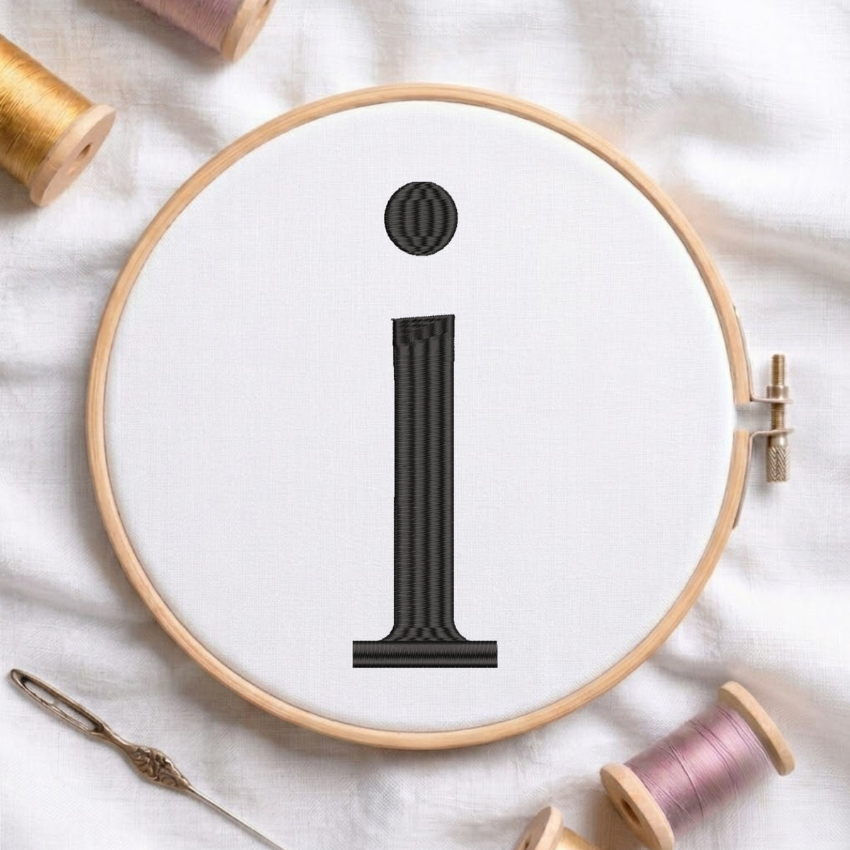 A wooden embroidery hoop holding white fabric with a black embroidered lowercase letter 'i'. Spools of thread and a needle are arranged around it on a white fabric background.