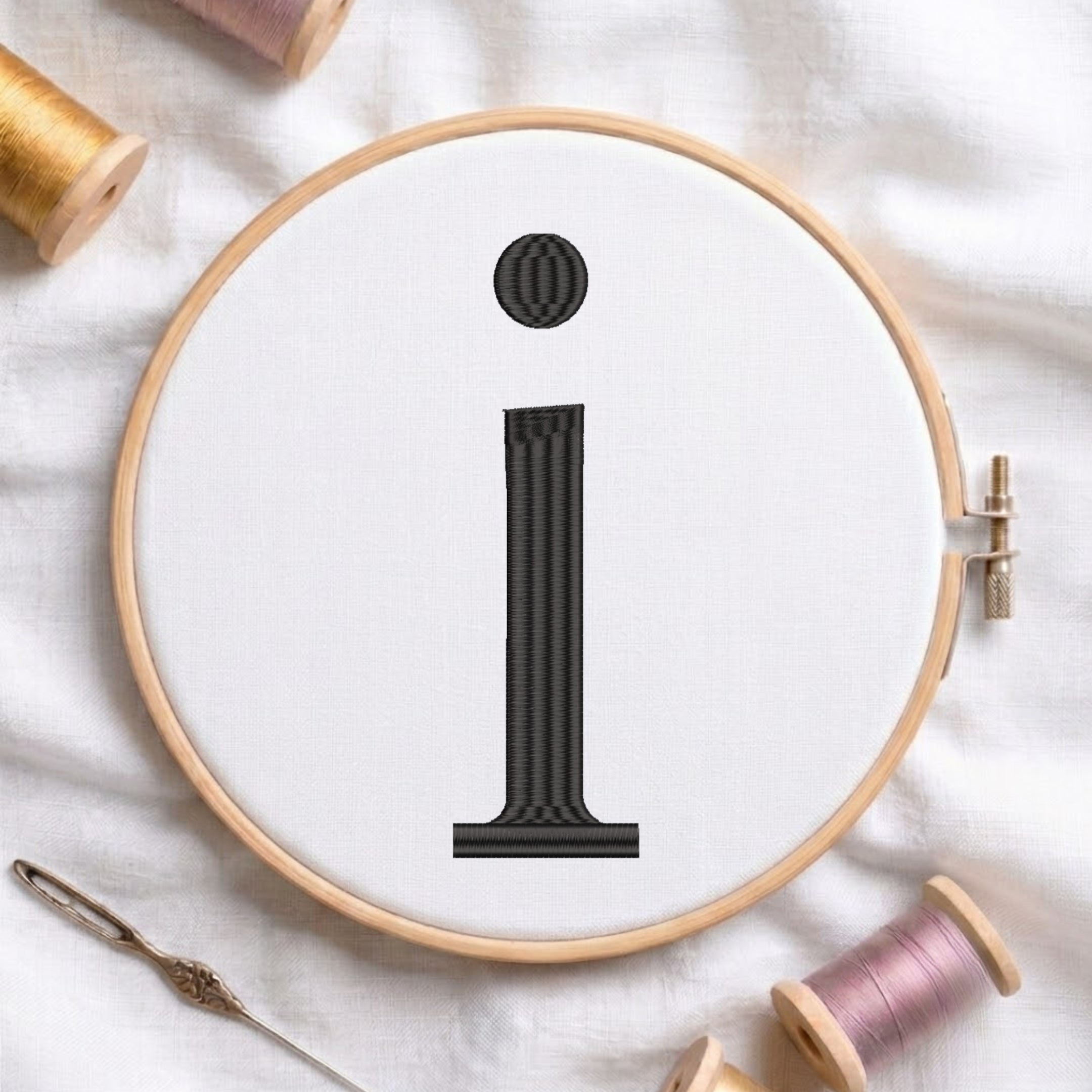 A wooden embroidery hoop holding white fabric with a black embroidered lowercase letter 'i'. Spools of thread and a needle are arranged around it on a white fabric background.