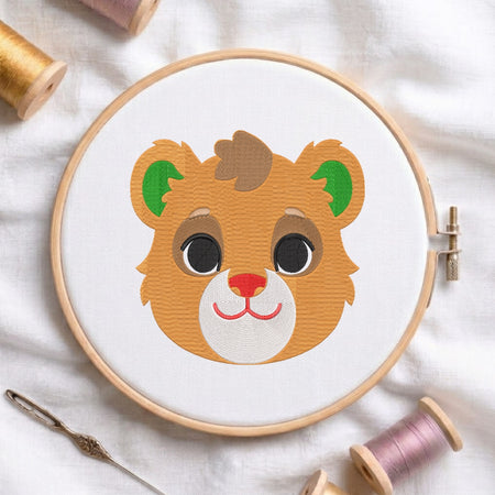 A circular embroidery hoop holding white fabric with a stylized cartoon bear face in orange, brown, green, black, and pink stitching. The hoop is photographed on a white fabric surface with spools of thread and a needle around it.