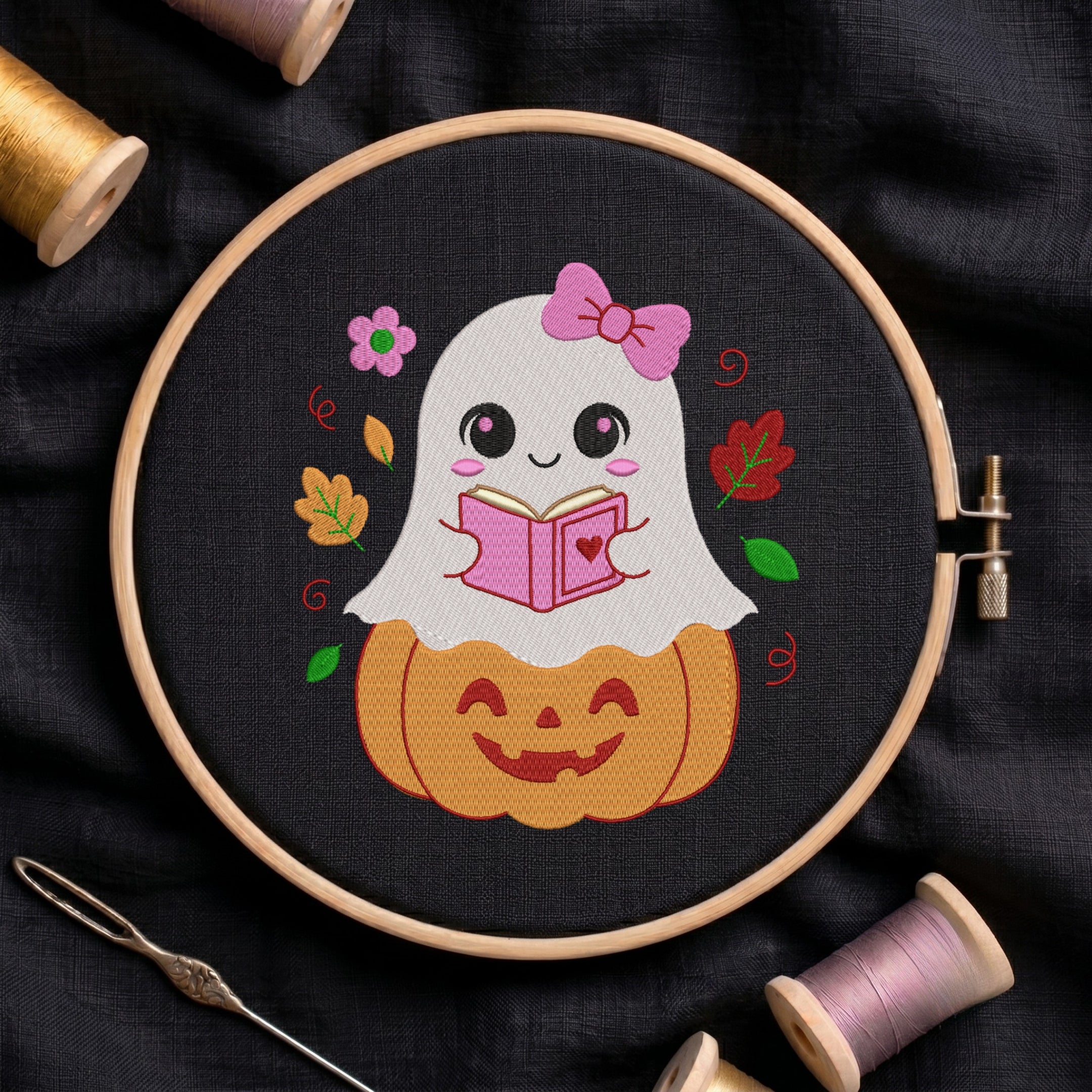 An embroidered design of a cute little ghost wearing a pink bow, sitting on a carved jack-o'-lantern pumpkin while reading a pink book, stitched inside a wooden hoop on black fabric with autumn leaves and sewing threads.