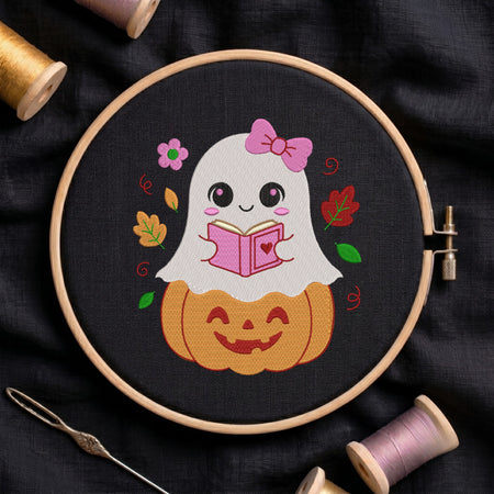 An embroidered design of a cute little ghost wearing a pink bow, sitting on a carved jack-o'-lantern pumpkin while reading a pink book, stitched inside a wooden hoop on black fabric with autumn leaves and sewing threads.