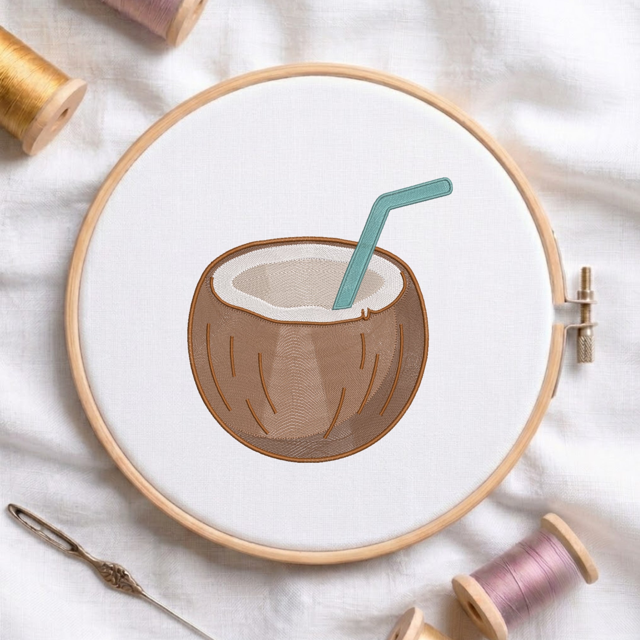 A wooden embroidery hoop holds white fabric with a stitched illustration of a brown coconut and a teal straw. Spools of thread and a needle are arranged around it on a white fabric background.