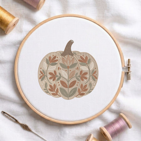 Floral-patterned pumpkin machine embroidery design displayed in a wooden hoop on white fabric with thread spools and a needle.
