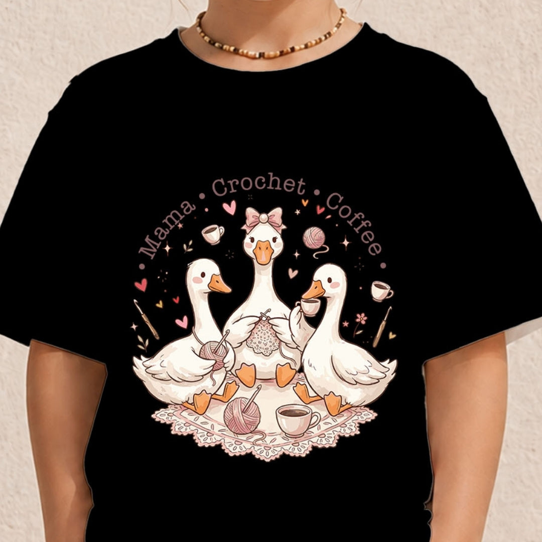 Close-up of a person wearing a black graphic t-shirt printed with a circular whimsical design of crocheting ducks, yarn balls, and coffee cups.