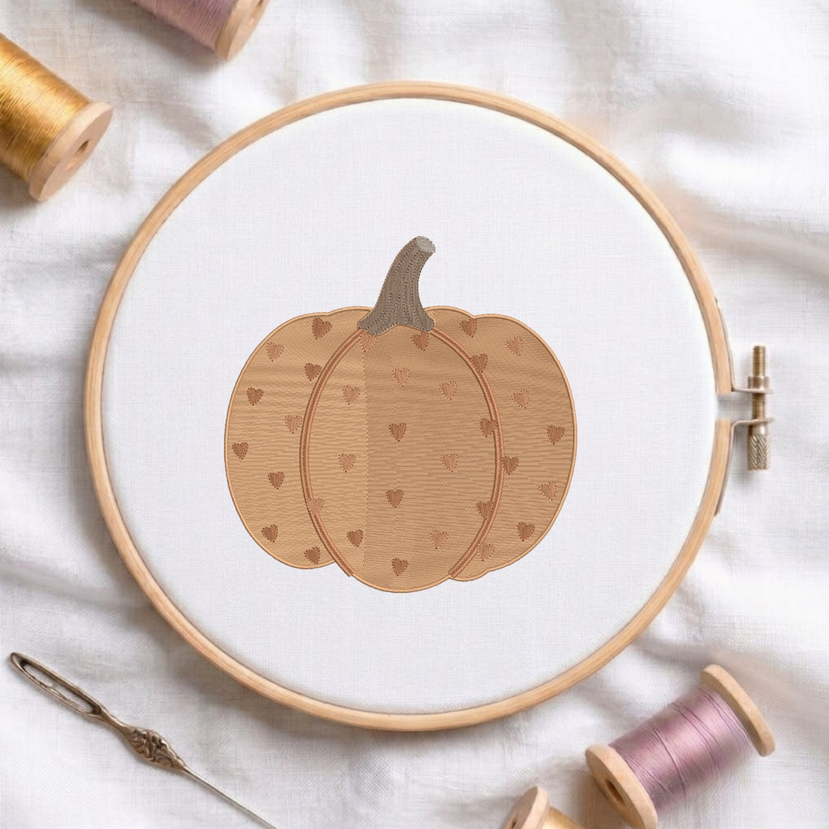 A circular embroidery hoop holding white fabric with a stitched pumpkin design in warm tan and brown tones with small heart motifs. The hoop is styled on white fabric with spools of thread and a needle around it.