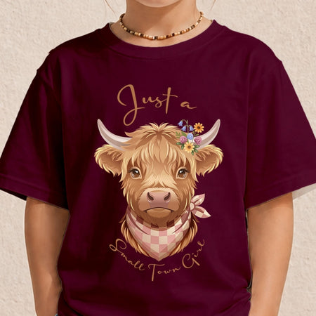 A person wearing a dark burgundy t-shirt with a graphic of a fluffy light-brown Highland cow wearing a pink checkered bandana and a flower crown, with the text 'Just a Small Town Girl' in gold script.