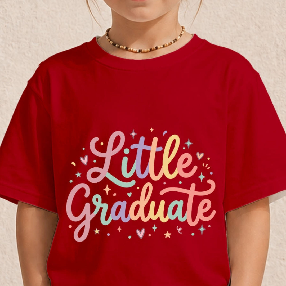 A child wearing a red short-sleeve T-shirt with a colorful pastel graphic that reads 'Little Graduate' in cursive lettering with stars and hearts, photographed against a light textured wall.