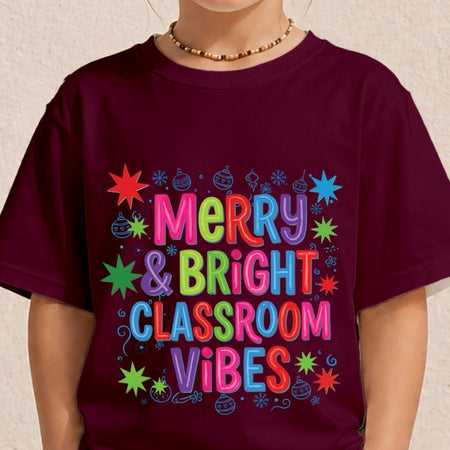A person wearing a dark burgundy t-shirt with a colorful graphic that reads 'Merry & Bright Classroom Vibes' in multicolored bubble letters surrounded by festive star and ornament illustrations.