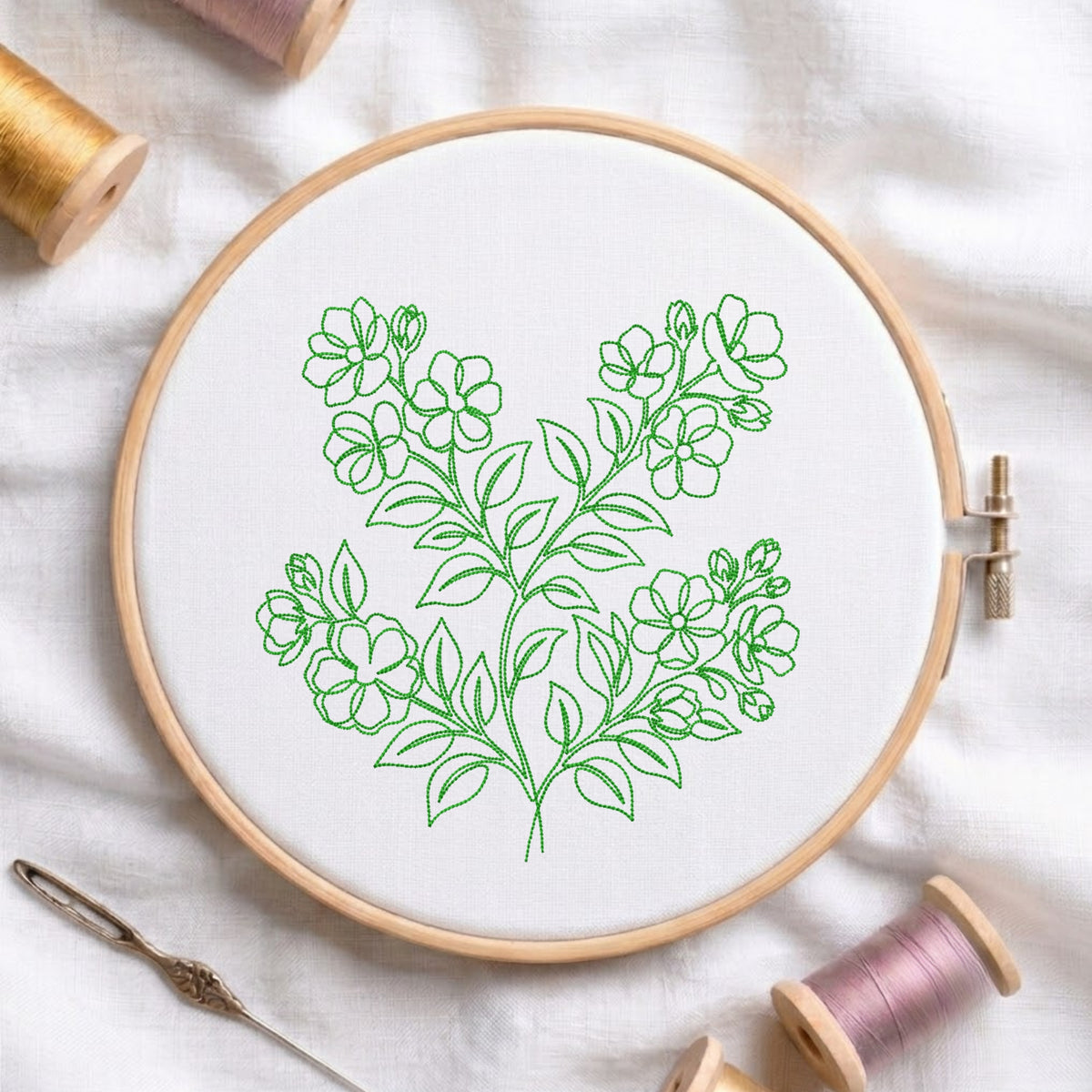 A wooden embroidery hoop holding white fabric with a green floral line design, surrounded by spools of thread and a needle on a white fabric background.