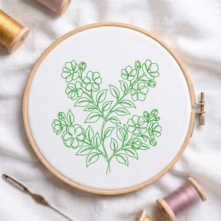A wooden embroidery hoop holding white fabric with a green floral line design, surrounded by spools of thread and a needle on a white fabric background.
