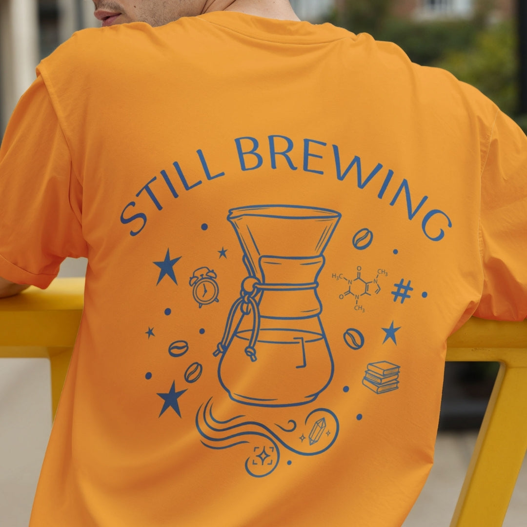 Rear view of a person wearing an orange t-shirt printed with a large blue "Still Brewing" pour-over coffee maker and caffeine molecule illustration across the back.