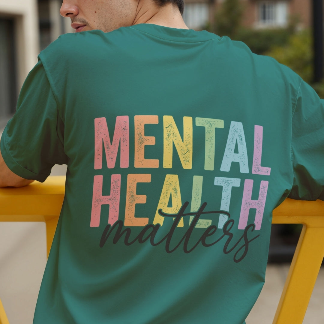 A man wearing a teal green t-shirt with a colorful graphic on the back that reads 'MENTAL HEALTH matters' in a distressed rainbow font and dark cursive script, leaning on a yellow railing in an outdoor setting.