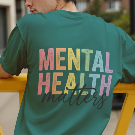 A man wearing a teal green t-shirt with a colorful graphic on the back that reads 'MENTAL HEALTH matters' in a distressed rainbow font and dark cursive script, leaning on a yellow railing in an outdoor setting.