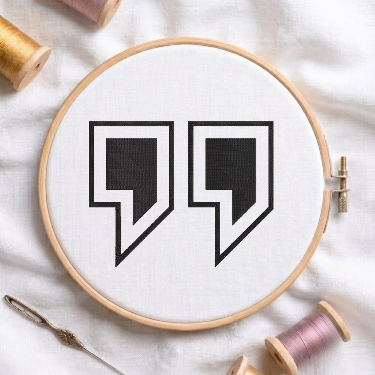 A circular embroidery hoop holding white fabric with a black quote-style symbol stitched in the center. The hoop is styled on a white fabric surface with spools of thread and a needle around it.