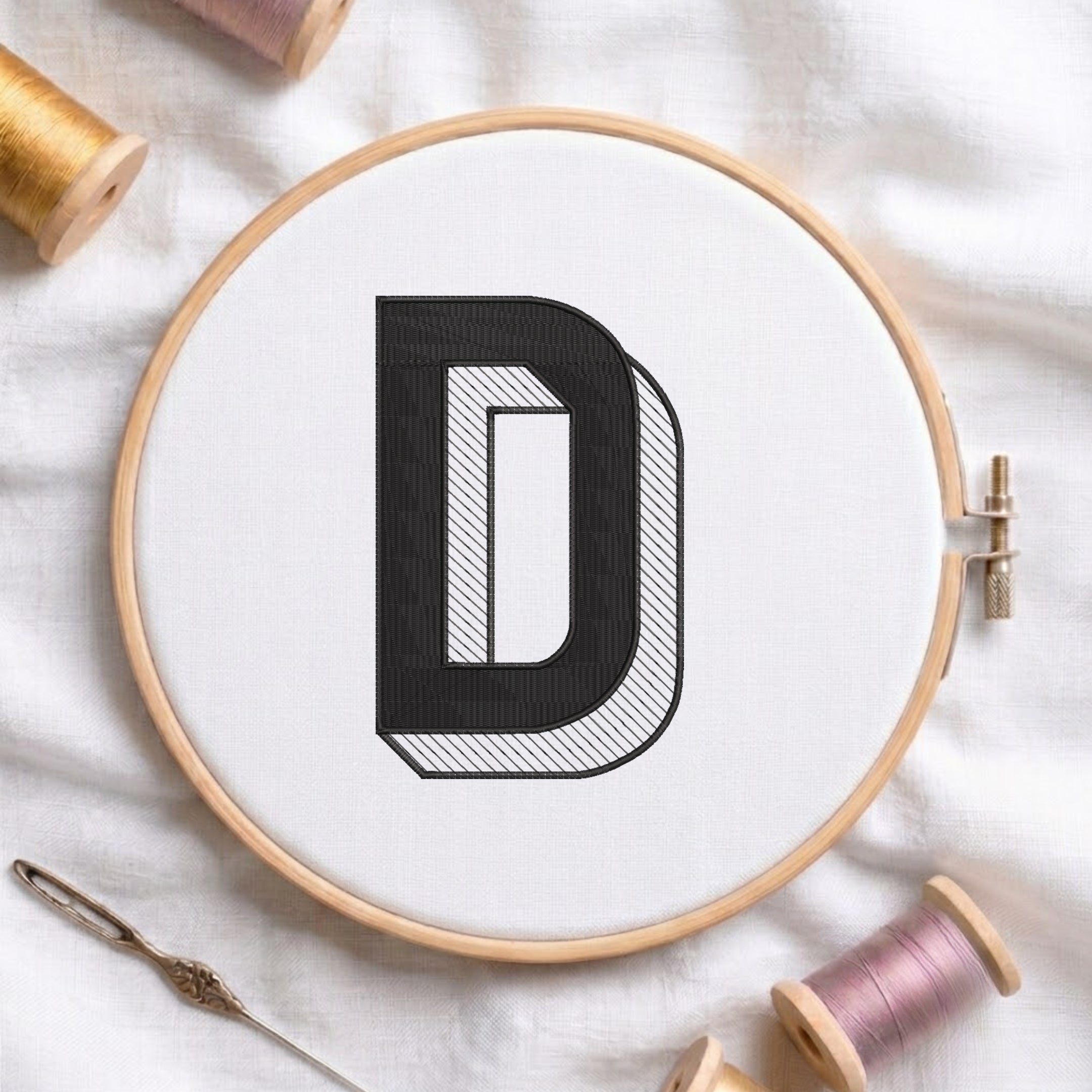 A wooden embroidery hoop holding white fabric with a large black embroidered letter D, styled on a white cloth with spools of thread and a needle around it.