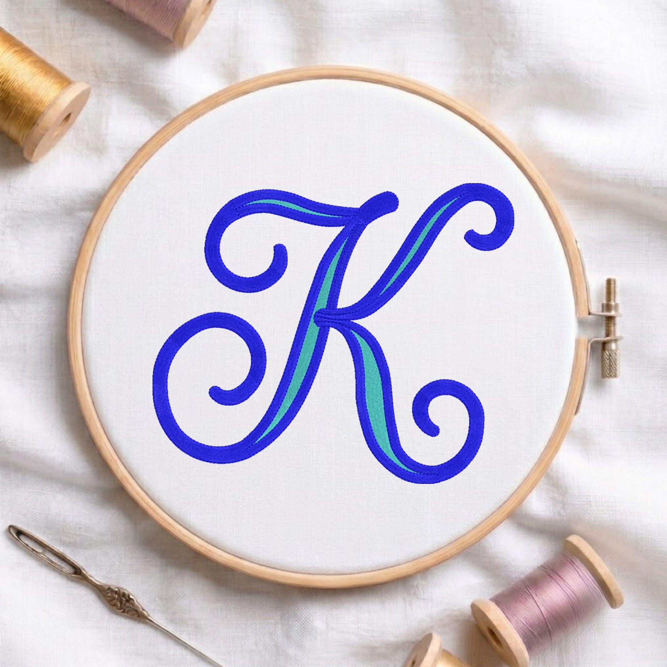 A wooden embroidery hoop holding white fabric with a large decorative letter K in blue and teal, surrounded by spools of thread and a needle on a white fabric background.