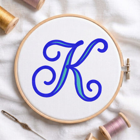 A wooden embroidery hoop holding white fabric with a large decorative letter K in blue and teal, surrounded by spools of thread and a needle on a white fabric background.