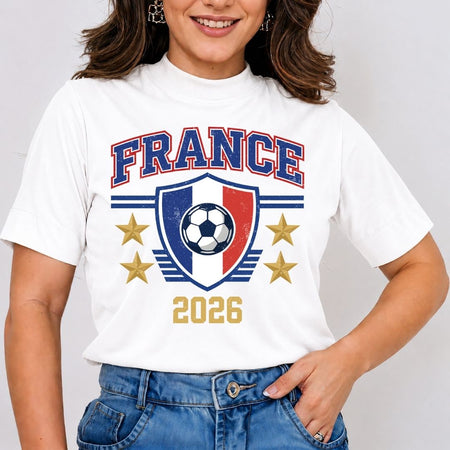 A woman smiling and wearing a white mock neck t-shirt featuring a retro France soccer graphic with blue and red crest bars, gold stars, and the year 2026.