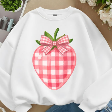 A white crewneck sweatshirt featuring a large pink and red gingham strawberry graphic with green leaves and a matching pink gingham bow, laid flat on a wooden surface with lifestyle props.