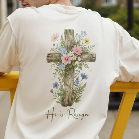 A person wearing a white t-shirt with a large floral cross graphic and the text "He is Resign" printed on the back.