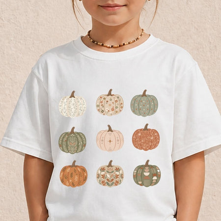 A child wearing a white short-sleeve T-shirt with a graphic of nine patterned pumpkins in muted autumn colors, photographed against a light textured wall.