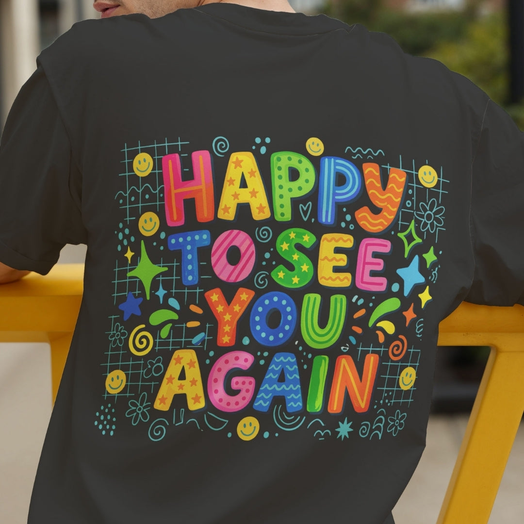 A person wearing a dark charcoal gray t-shirt, shown from the back. The shirt features a large, colorful retro-style graphic that reads 'HAPPY TO SEE YOU AGAIN' in playful, hand-drawn letters with doodles of stars, smiley faces, and lines.