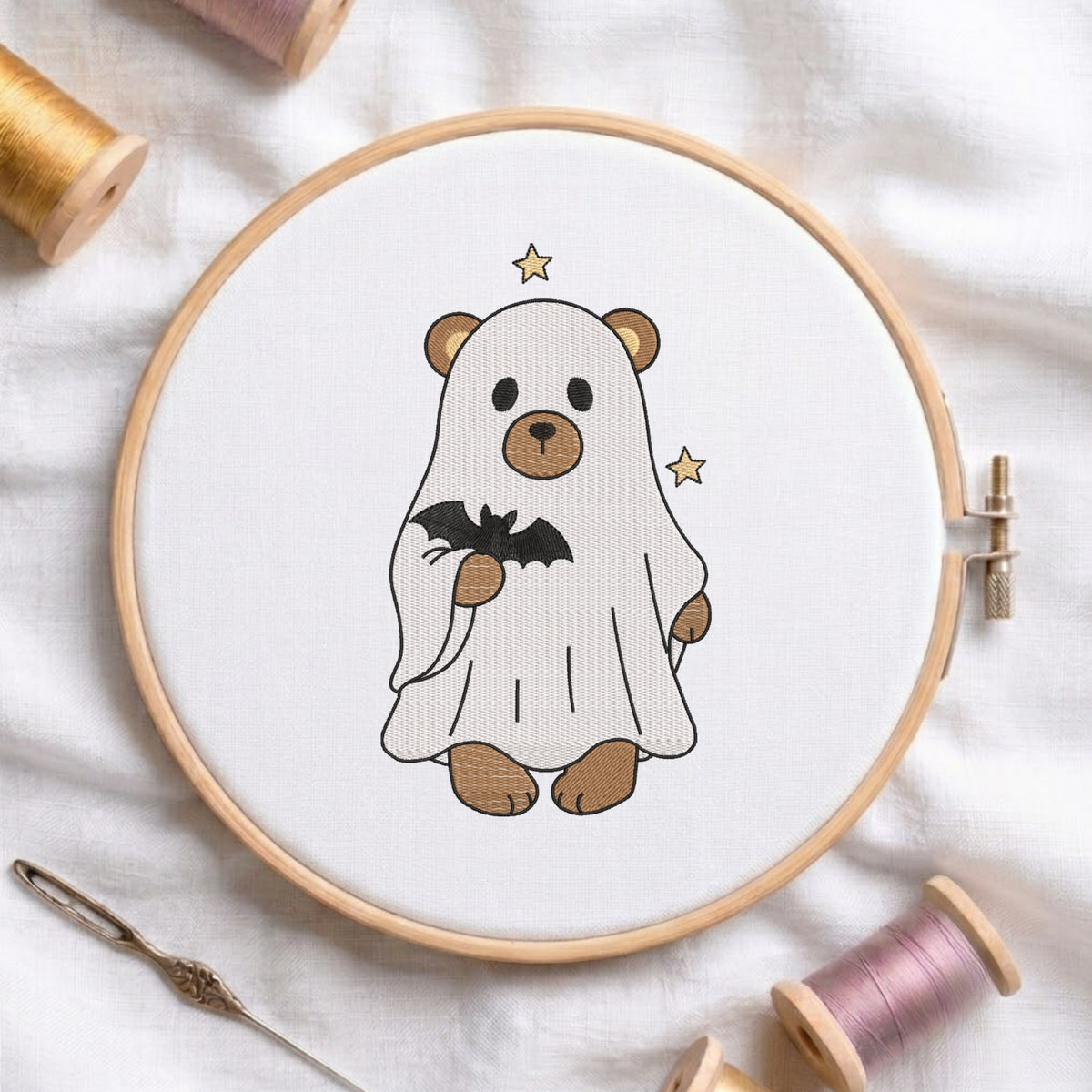 An embroidered brown bear wearing a white ghost sheet and holding a black bat, stitched inside a wooden hoop on white fabric with sewing threads.