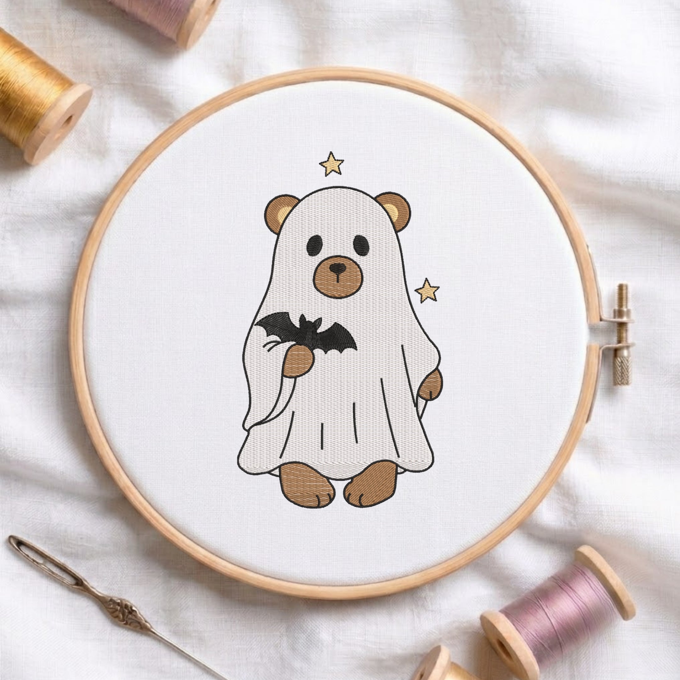 An embroidered brown bear wearing a white ghost sheet and holding a black bat, stitched inside a wooden hoop on white fabric with sewing threads.