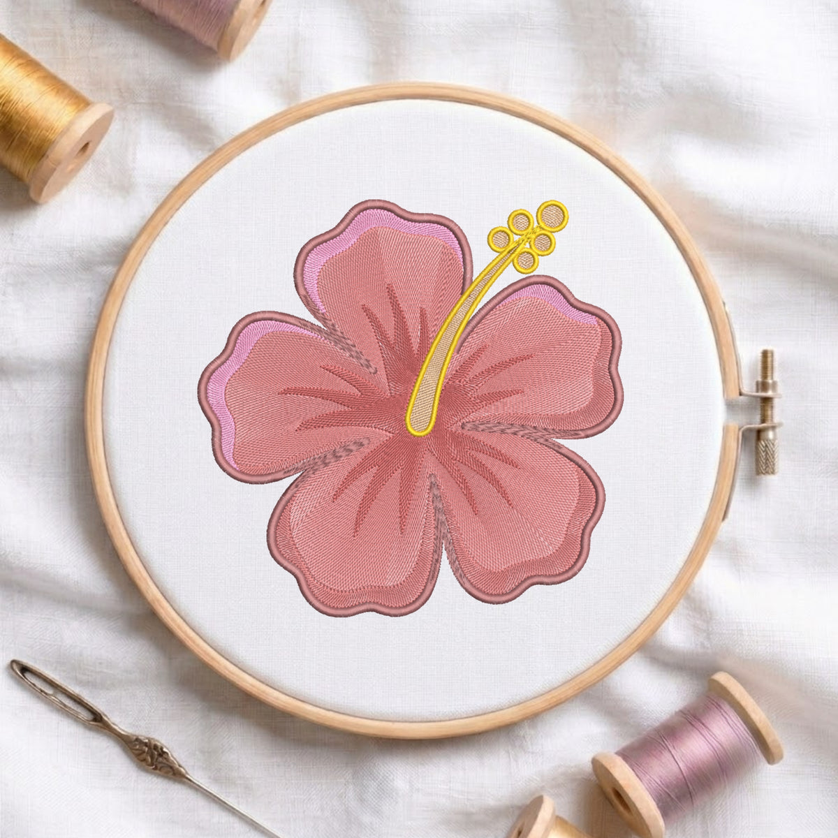 A circular embroidery hoop holding white fabric with a stitched pink hibiscus flower design outlined in brown, surrounded by spools of thread and a needle on a white fabric background.
