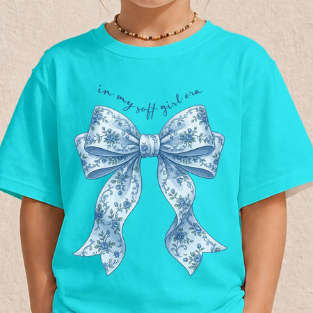 A person wearing a bright turquoise t-shirt featuring a blue floral bow graphic and the text "in my soft girl era."