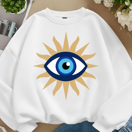 A white crewneck sweatshirt featuring a large graphic of an evil eye surrounded by a gold sunburst, laid flat on a wooden surface with decorative plants and flowers in the background.