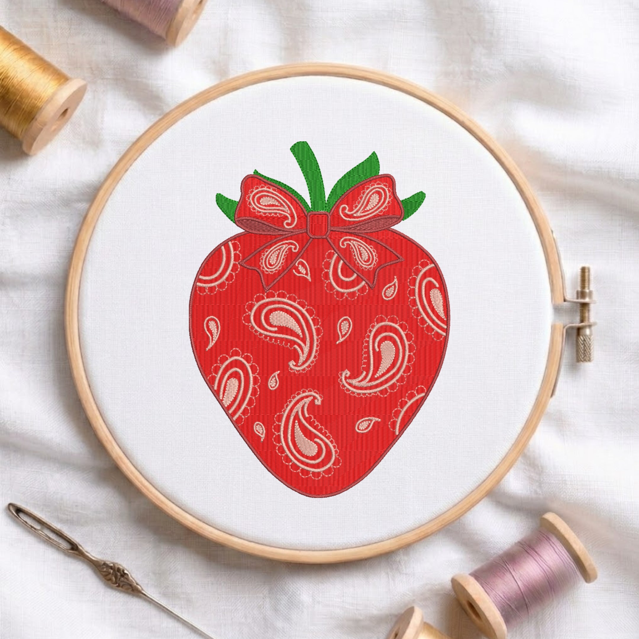 A close-up of a red strawberry with a paisley pattern and bow embroidery design, stitched on white fabric inside a wooden embroidery hoop surrounded by spools of thread and a needle.