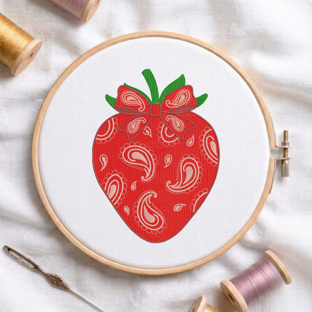 A close-up of a red strawberry with a paisley pattern and bow embroidery design, stitched on white fabric inside a wooden embroidery hoop surrounded by spools of thread and a needle.