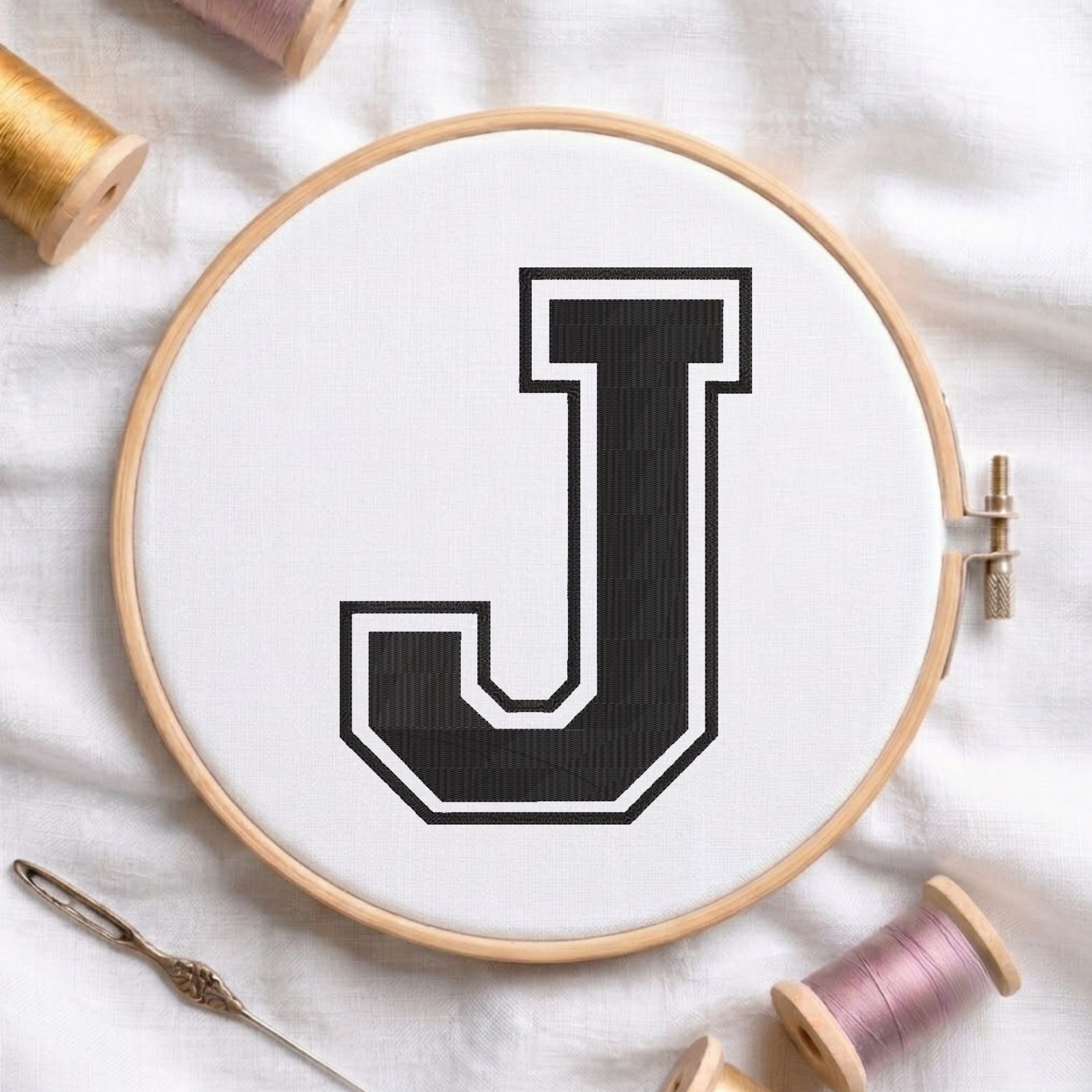 Collegiate style letter J machine embroidery design in black with white outline, displayed inside a wooden embroidery hoop with golden and pink thread spools.