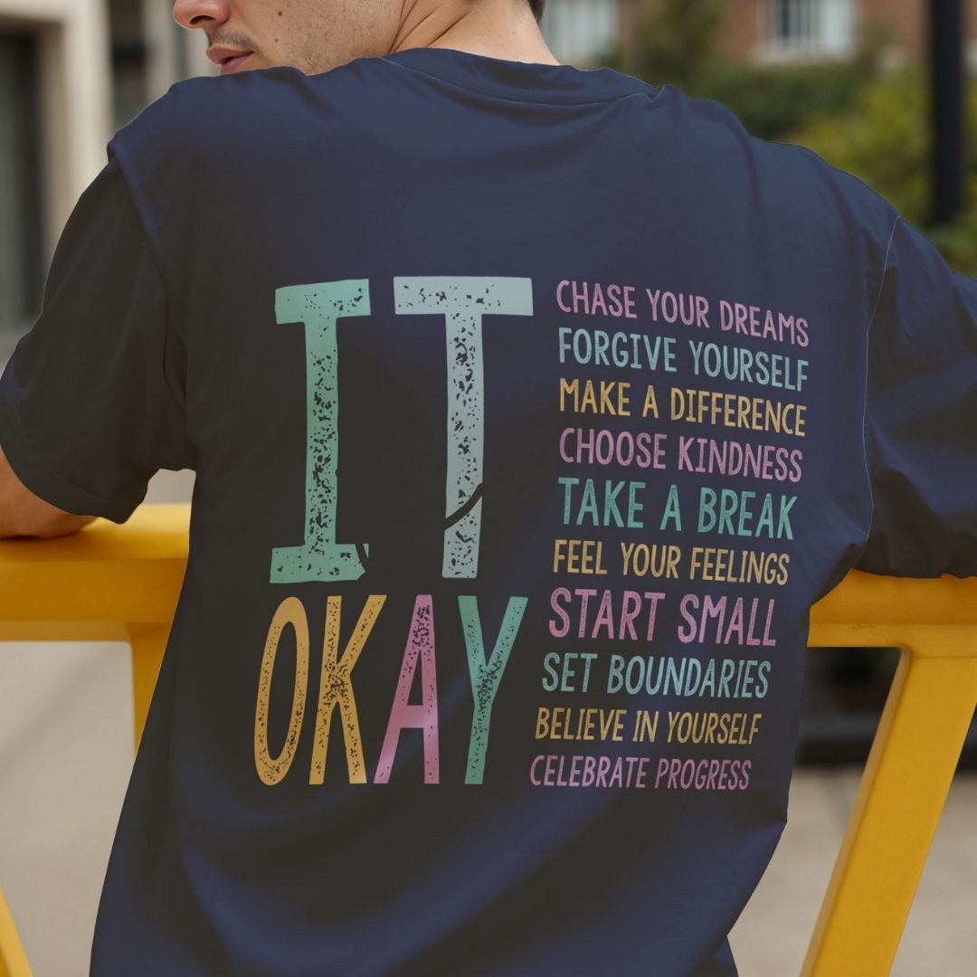 A man wearing a navy blue t-shirt from the back, featuring a colorful graphic with the words 'IT OKAY' and a list of positive affirmations in pastel colors.