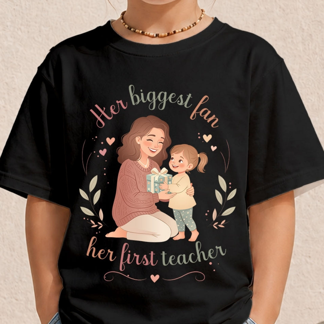 Close-up of a child wearing a black crewneck t-shirt with a sweet illustration of a mother and daughter surrounded by hearts and the text Her biggest fan, her first teacher.