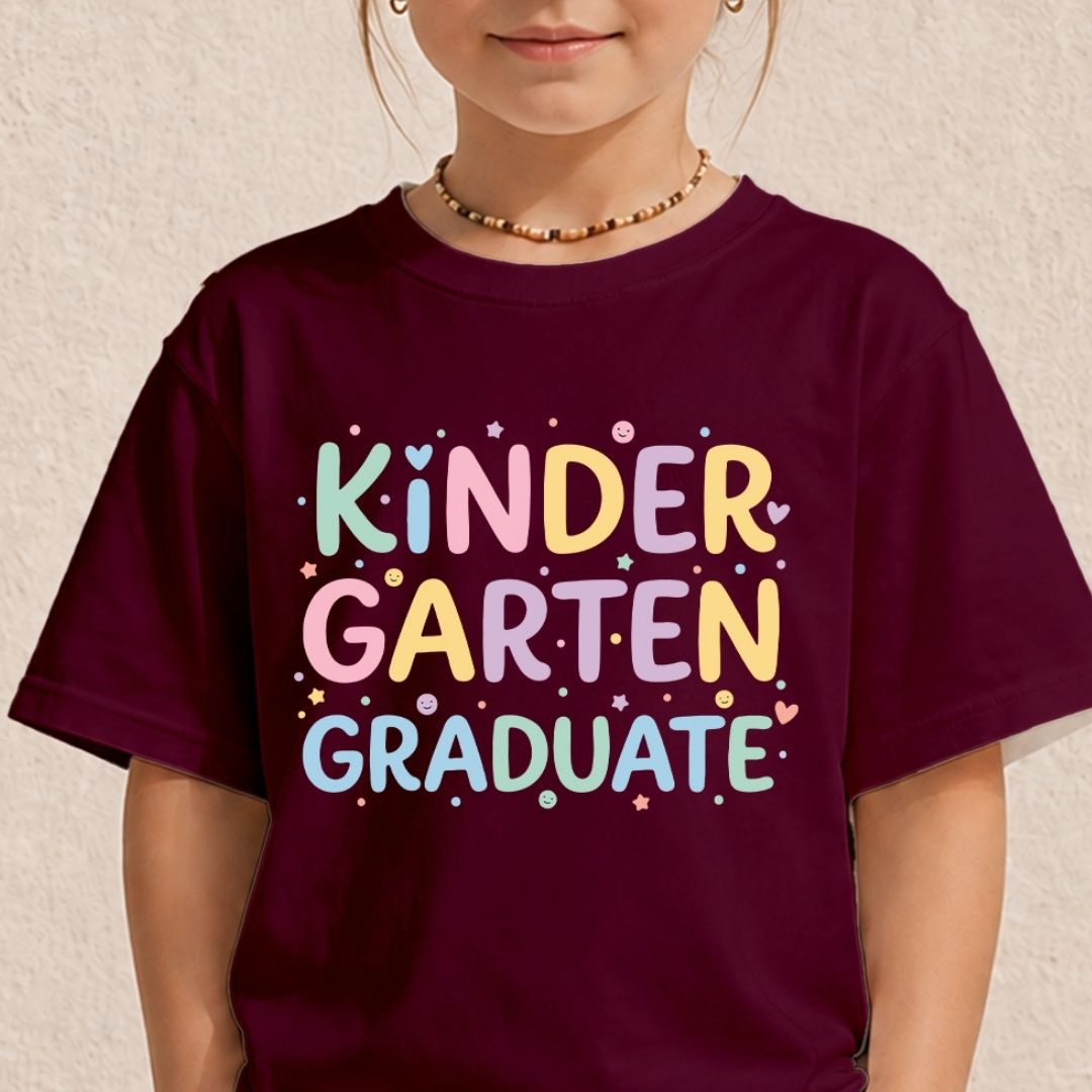 A child wearing a dark maroon t-shirt with a colorful graphic that reads 'KINDER GARTEN GRADUATE' in pastel letters with small star and heart accents, set against a textured beige wall.
