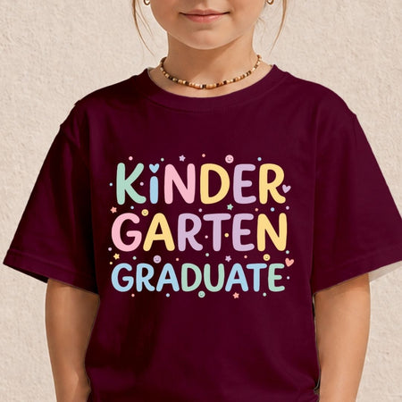 A child wearing a dark maroon t-shirt with a colorful graphic that reads 'KINDER GARTEN GRADUATE' in pastel letters with small star and heart accents, set against a textured beige wall.