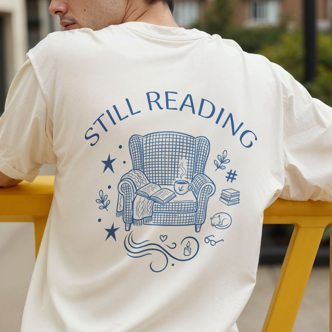 Rear view of a person wearing a cream-colored t-shirt printed with a large blue "Still Reading" armchair and book illustration across the back.