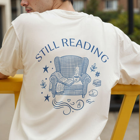 Rear view of a person wearing a cream-colored t-shirt printed with a large blue "Still Reading" armchair and book illustration across the back.