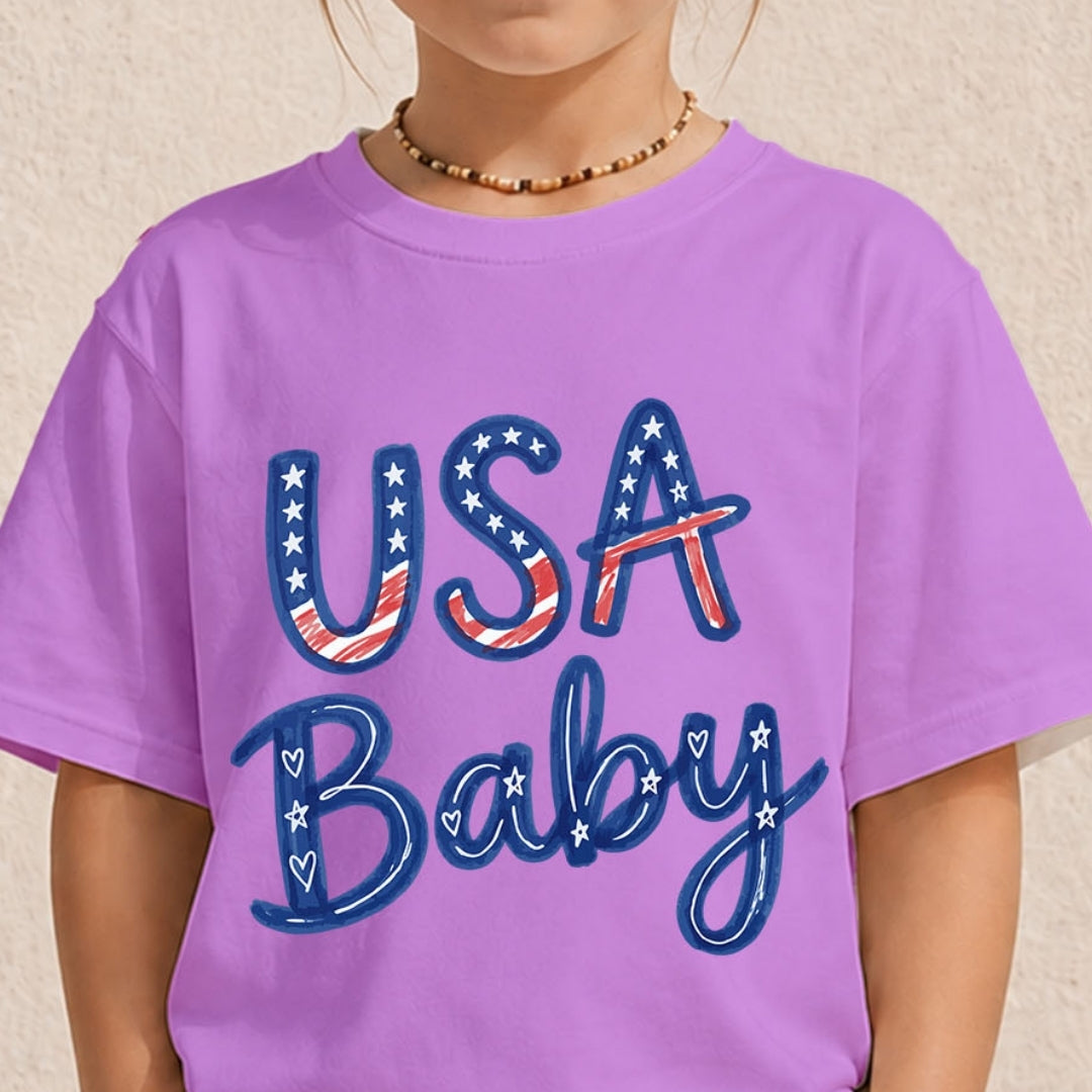A child wearing a lavender purple t-shirt with a patriotic graphic that reads 'USA Baby' in blue, red, and white. The child is also wearing a beaded necklace, set against a neutral textured beige background.