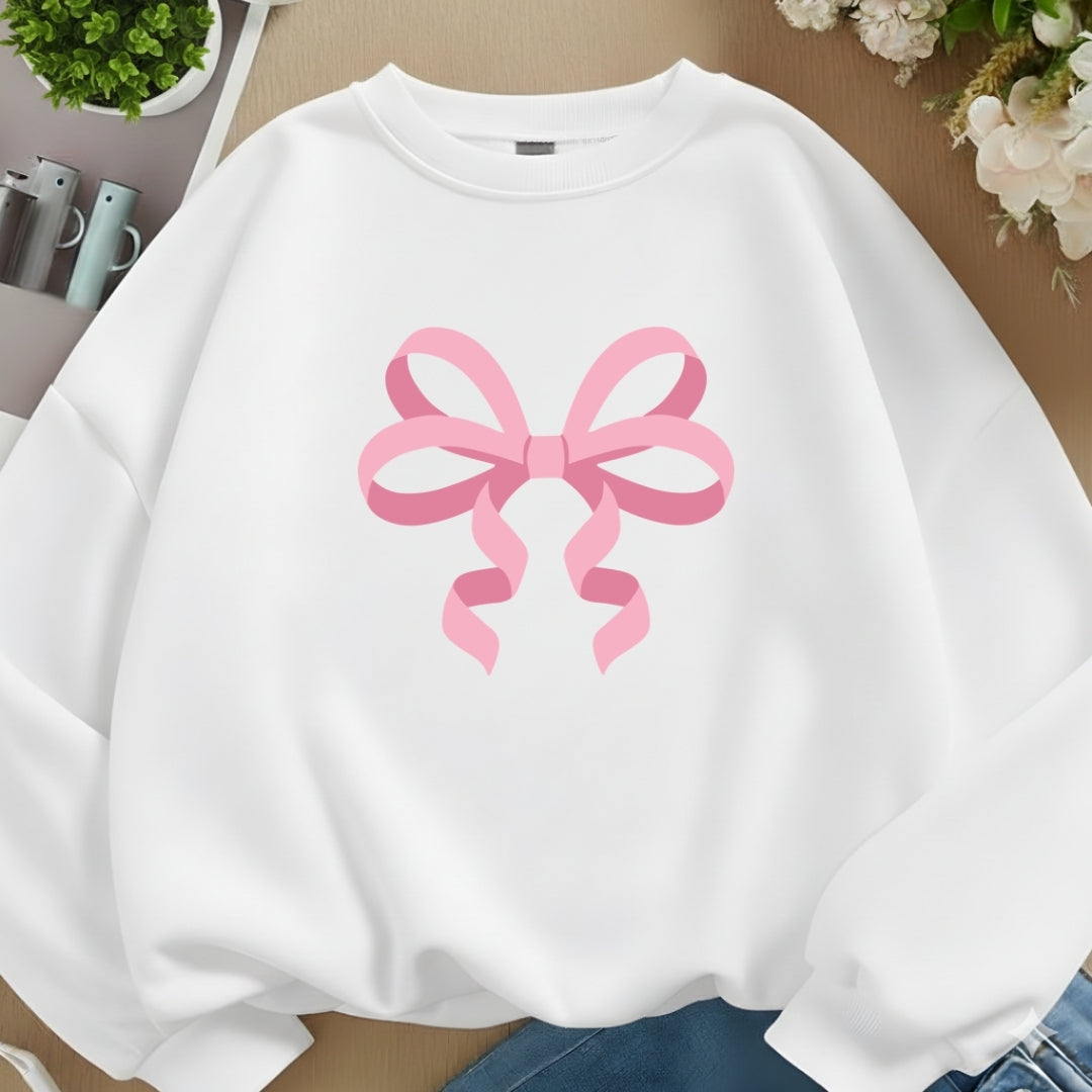 A white crewneck sweatshirt featuring a large pink ribbon bow graphic printed on the chest, styled in a flat-lay arrangement with blue jeans, flowers, and small plants.