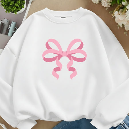 A white crewneck sweatshirt featuring a large pink ribbon bow graphic printed on the chest, styled in a flat-lay arrangement with blue jeans, flowers, and small plants.