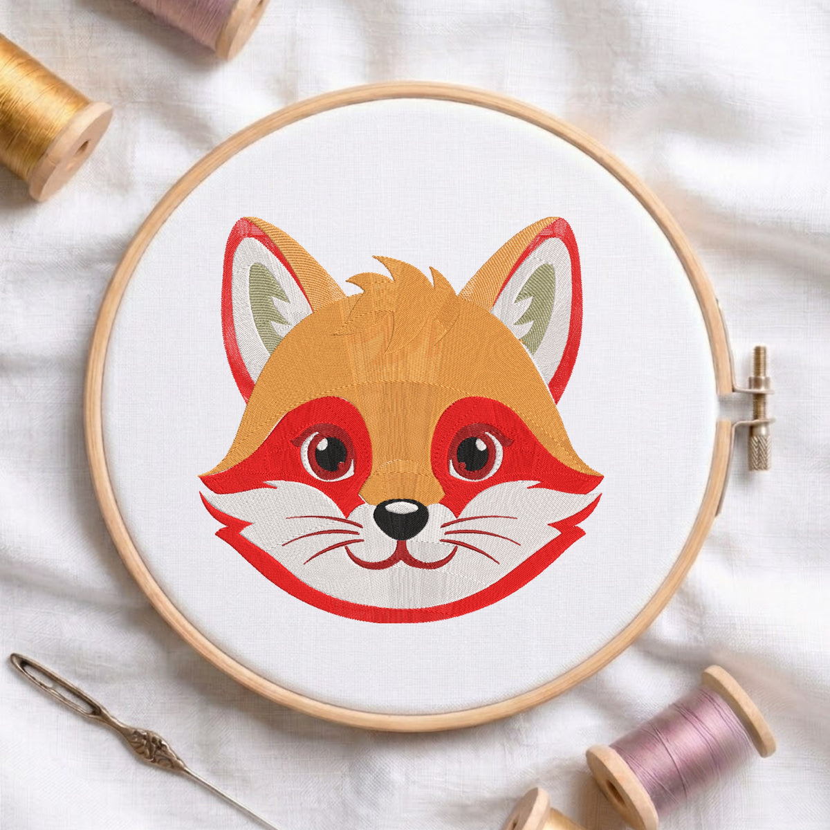A circular embroidery hoop holding white fabric with a stylized fox face design in orange, red, white, and black. The hoop is photographed on a white fabric background with spools of thread and a needle around it.