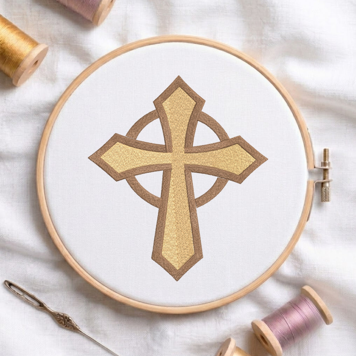 An embroidery hoop on a white cloth displays a finished machine embroidery design of a textured gold-yellow Celtic cross with a darker brown outline and intersecting ring, flanked by spools of thread and a needle.