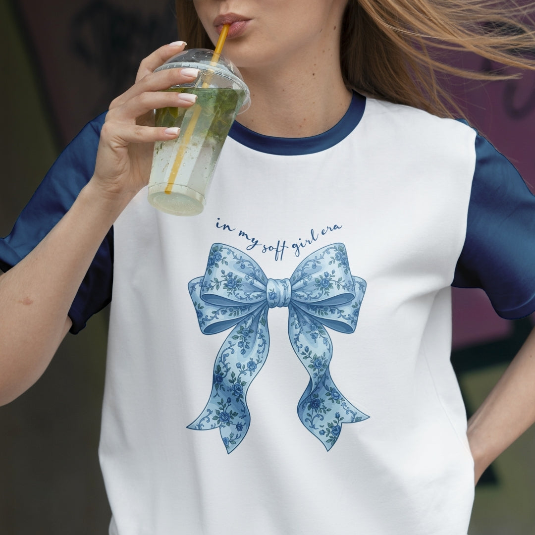 Person wearing a white t-shirt with a blue bow design and text, holding a drink.