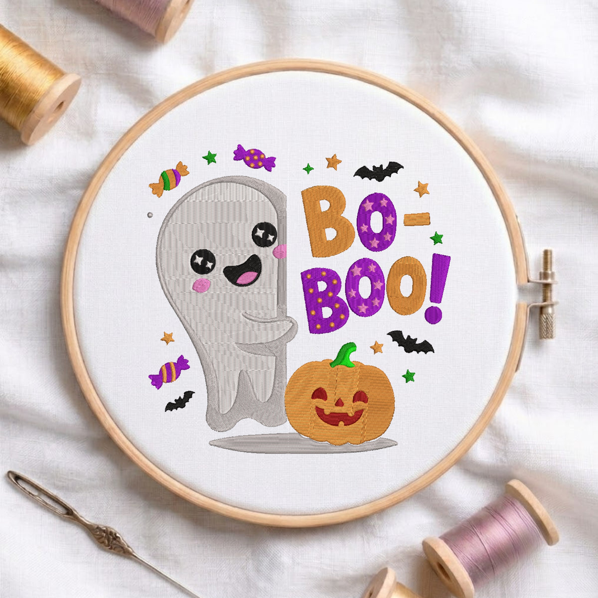 A circular embroidery hoop on white fabric, featuring a Halloween-themed design with a smiling gray ghost, a jack-o'-lantern pumpkin, colorful candy motifs, bats, stars, and the words “BOO-BOO!” in orange and purple. Spools of thread and a needle are arranged around the hoop.